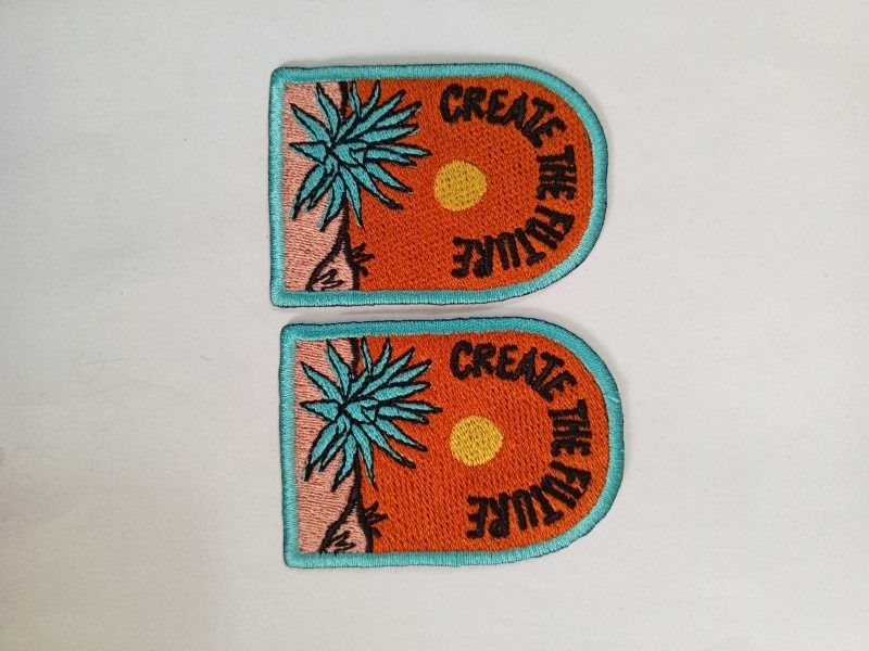 Bring the artwork. Two orange and turquoise patches with a flower and Creative lettering, photographed on pale wood. Liberty Digitizing design with black, white and gold typography.