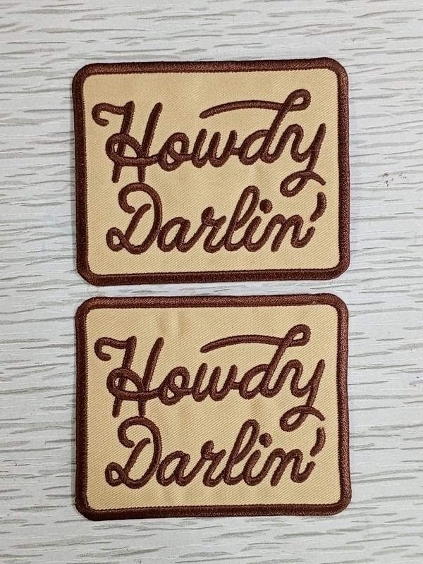 A little character. A lot of thread. Two cream patches with brown Howdy Darlin lettering photographed on pale wood. Liberty Digitizing design with black, white and gold typography.