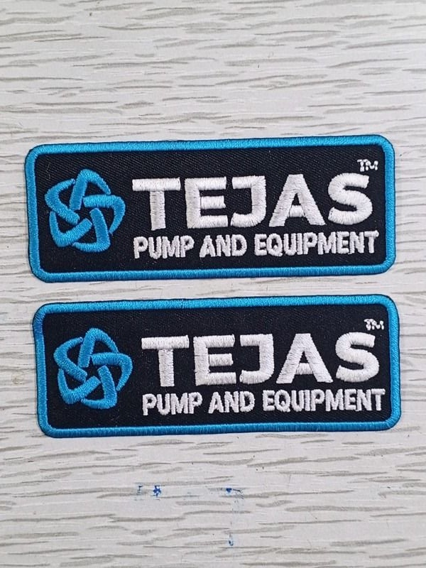 Your logo. On the job. Two Tejas Pump and Equipment logo patches with blue borders and white lettering, photographed on pale wood. Liberty Digitizing design with black, white and gold typography.